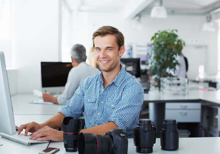 a man wearing a blue dress shirt sitting at a computer with camera equipment on the desk.