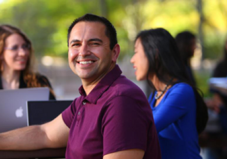 Image of a male student smiling sitting by computer with other students in the room